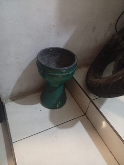 The scene is an interior corner in Margahayu Tengah, Indonesia. A dark green, goblet-shaped object, approximately 30 cm in height, rests centrally on a light-colored tiled floor. The object features a wide, dark-textured opening at the top, a tapering body with two horizontal bands, and a flared base showing minor white scuff marks. To the right, partially visible against the wall, is a segment of a dark, worn object, consistent with a tire. The floor is composed of square, light-colored tiles with dark gray grout lines. The background wall is off-white and shows surface texture and minor discoloration, particularly at its junction with the floor. A distinct shadow from the green object extends onto both the wall and the floor, indicating a light source positioned above and to the left. No individuals or active interactions are present.