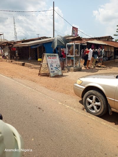 The image shows a street scene in Okinni, Nigeria, taken from a car parked along the side of the road. The road is paved and has a yellow dividing line. To the right of the road is a sidewalk with a dirt surface. There are a number of small buildings on the right side of the road. One building has a sign that reads "OPEYEMI FISH DEPOT." There are several mannequins in front of the building, wearing clothing. A car is parked on the right side of the road. The sky is clear and blue with a few white clouds.
