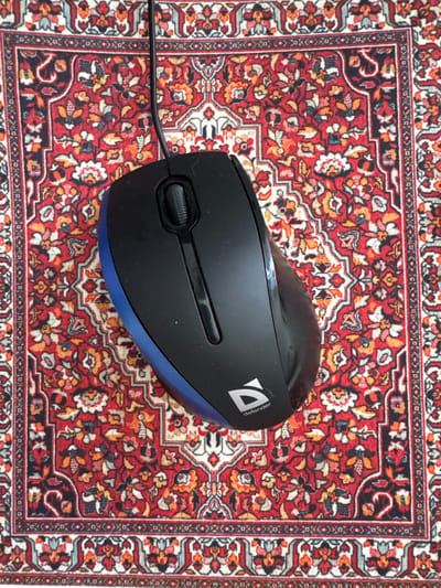 A black computer mouse with a blue trim is positioned on a red oriental rug with intricate patterns. A black wire extends from the top of the mouse. The rug is adorned with floral motifs in shades of red, white, blue, and orange. The mouse has a scroll wheel and a logo that reads "defender". The image is taken from a top-down perspective. The scene is indoors, on a flat surface. There are no people or animals visible in the image. The specific location in Боголюбово, Russia is not discernible from the provided image.