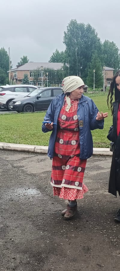 The image shows two women standing outside on a cloudy day, likely in Старые Быги, Russia. The woman in the foreground is wearing a traditional outfit, including a light-colored headscarf and a red and white checkered dress with decorative white appliques. She has a blue jacket on and is holding a blue object in her right hand. Her expression suggests she is engaged in conversation or explaining something, with her hands gesturing outwards. The second woman, partially visible on the right, is wearing a black coat.
Behind the women, a grassy area is visible, bordered by a curb and a paved surface that appears wet from rain, indicated by puddles on the ground. In the background, several cars are parked, including a white SUV and a dark sedan. Further back, there is a brick building with a light-colored roof, and tall green trees line the area. The overcast sky suggests it is either morning or afternoon, with a damp and cool atmosphere. The overall scene depicts an informal outdoor gathering or moment on a possibly rainy day. No visible text is present in the image.