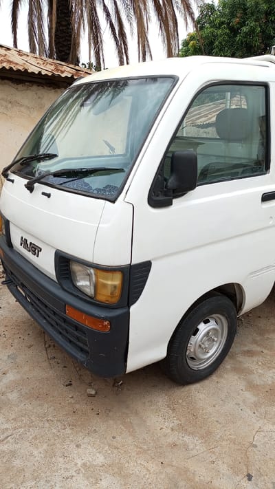 A white Daihatsu Hijet truck is parked on a cracked concrete surface in an outdoor environment, likely in Kawo, Nigeria. The front end of the vehicle is visible, with a black front bumper, yellowed headlights, and the word "HIJET" displayed. A tire and wheel are visible on the right side of the image. A building with a rusty corrugated iron roof is in the background on the left side of the image, and trees are visible in the background on the right side. Palm tree fronds overhang the scene from above. No people are visible.