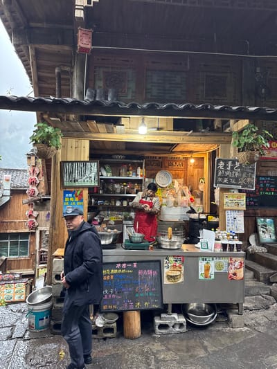The image depicts a street food stall in Gongtan, China, under overcast conditions. In the foreground, a middle-aged Asian male stands on a wet stone-paved ground, facing slightly left. He wears a dark baseball cap, a dark puffy jacket, dark trousers, and dark shoes, with his left hand near his waist.

Behind him, within an open-fronted traditional wooden structure, a middle-aged Asian female vendor in a red apron over a light shirt is actively preparing food at a stainless steel counter. She holds a small, round item in her hands, possibly dough. On the counter, there are multiple metal bowls, a pot on a small cooktop, and a deep fryer basket containing fried items. Shelves behind her are stocked with jars and bottles of ingredients. A large white decorative plate is mounted on the back wall, which also features ornate wooden panels.

Menu items are displayed on several black chalkboards and white signs. A left chalkboard details "Iced Slush Summer" options like Osmanthus Rice Wine Iced Jelly, while a right chalkboard advertises "Old Craft" and "Fresh Meat Pancake." A red sign to the left promotes "Freshly Brewed Sour Plum Drink." A bare lightbulb illuminates the stall interior.

To the left of the stall, another traditional wooden building with a tiled roof and a vertical red-character sign is visible. A woven basket with greenery hangs near the stall's entrance. The ground is wet and reflective, with concrete blocks supporting the counter and other items. In the distant background, hazy mountains are visible under an overcast sky.