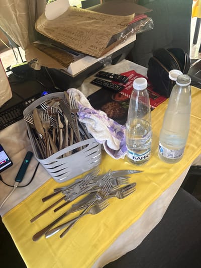 The image depicts an indoor scene in the city of Hrishteni, Bulgaria, showcasing a table laden with various objects. The table is covered by a yellow tablecloth, beneath which a white layer is visible.
In the background, a stack of brown paper sheets, printed with patterns such as "BON APPETIT," "CAFE & CAFE," "Coffee," and "BOULANGERIE," rests near a crumpled white curtain. To the right, a black quilted backpack or purse with golden zippers is open. Two black remote controls are positioned on a red placemat that features an image of a cooked patty and partially visible Cyrillic text including "250 м." and "към."
On the left side of the table, a black keyboard is partially visible, alongside a smartphone connected via a black cable and a dark green/blue stick-like device with a green indicator light, which is also connected via a white USB cable.
A light grey plastic basket, filled with numerous silver-colored forks and knives, occupies the center-left. A white towel with purple stripes is draped over the side of this basket. In the foreground, additional silver-colored forks and knives are loosely spread on the yellow tablecloth.
To the right, two clear plastic water bottles are present. The left bottle, partially filled with water, has a white cap and a label reading "БАНКА" (BANKA) and "НАТУРАЛНА МИНЕРАЛНА ВОДА." The right bottle is mostly empty, its white cap slightly ajar, and it also features the "БАНКА" label with an indication of "1 LE" (1 Liter). No people are visible in the scene.