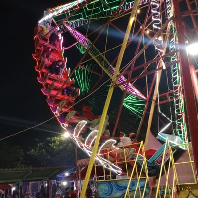 The image shows a Ferris wheel with people riding it. The Ferris wheel is located in Kecamatan Curug, Indonesia at the coordinates -6.262, 106.558. The wheel is brightly lit with multicolored LED lights. The Ferris wheel is made of metal and has a red and yellow color scheme. It appears to be a small Ferris wheel with a limited number of gondolas. The people on the Ferris wheel are smiling and appear to be having a good time. The Ferris wheel is situated in a public park or carnival setting. There are trees and other amusement rides in the background. There is a sign in the bottom right corner of the image that reads "2019-1-25". The image was taken at night, and the Ferris wheel is the only thing that is well lit. The image appears to be taken by a person standing on the ground looking up at the Ferris wheel.