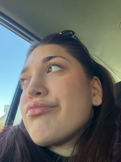 A young adult female is depicted in a close-up, angled portrait from within a vehicle. She has dark hair, partially pulled back and secured with a dark hair clip visible at the top of her head. Her complexion is light to medium, with brown eyes featuring long eyelashes, and well-defined eyebrows. Her lips are full and appear glossy, pursed into an expressive formation. Her gaze is directed upwards and to the viewer's right. A dark collar is visible at the base of her neck. The immediate environment includes a light-colored vehicle headliner and a dark window frame. Beyond the window on the left, a clear blue sky is visible above a faint, distant light-colored building or structure. The scene is located in Mutzig, France.