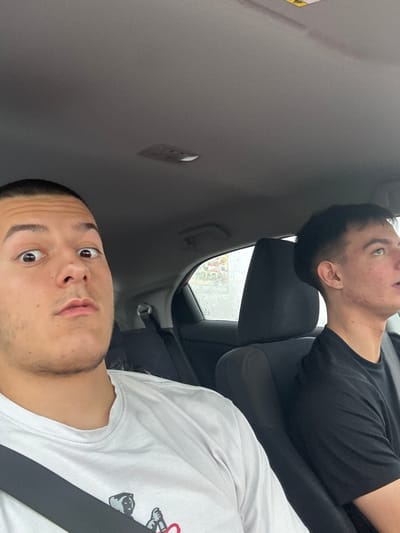 Two young men are sitting in the front seats of a car. The man in the foreground is wearing a white t-shirt and a seatbelt, looking directly at the camera with a surprised expression. The man in the background is wearing a black t-shirt and is looking upwards. The setting is indoors, likely inside a vehicle. Krustina, Bulgaria is not visibly identifiable in the image.
