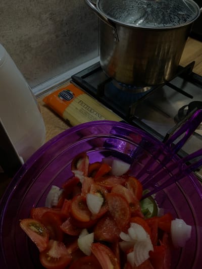 A stovetop with a pot of boiling water is visible, with a package of fettuccine pasta nearby. In the foreground, a purple bowl contains chopped tomatoes and onions, with purple serving utensils resting on the edge. The scene appears to be a kitchen in Rustavi, Georgia.
