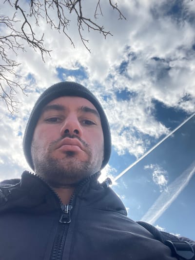 An adult male is depicted in an upward-angled outdoor selfie, taken in Lesidren, Bulgaria. The subject is positioned centrally, showing his upper torso, face, and head. He is wearing a dark knit beanie and a dark zippered jacket, with the prominent zipper and pull tab visible on his chest. He has dark eyebrows and short facial hair, with a neutral expression and slightly pursed lips. The background features a partly cloudy sky, displaying patches of blue amidst white clouds. Bare tree branches are visible in the upper left quadrant, silhouetted against the sky. A distinct, straight white contrail from an aircraft diagonally traverses the upper right portion of the sky, with another less defined contrail visible further left. The scene is brightly illuminated by natural light.