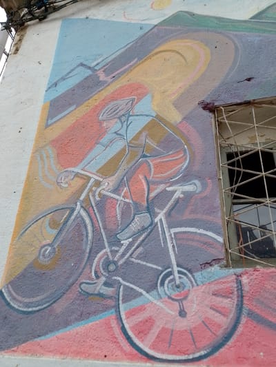 Mural depicting a cyclist riding on an uphill road, painted on an exterior wall in Municipio Tarija, Bolivia. The mural is rendered in a stylized, slightly abstract manner. The cyclist is wearing a helmet and cycling attire. The road is represented by colorful stripes of yellow, orange, purple, and pink. Mountains are depicted in the upper left corner. A barred window is visible on the right side of the mural. The wall is white and slightly textured.