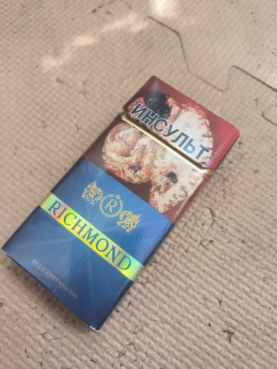 A close-up, overhead shot shows a cigarette pack lying on a textured, light brown surface, possibly a mat. The pack is dark blue with a holographic band that reads "RICHMOND" in yellow letters. Above the band, a graphic depicts a human brain, with Russian text reading "ИНСУЛЬТ" (stroke) in white letters at the top. The bottom of the pack has smaller text that reads "BLUE EDITION 100". The lighting casts shadows on the surface.