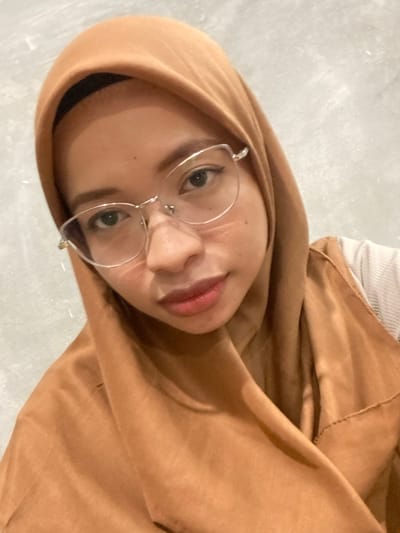 A woman wearing a brown hijab and glasses is captured in a close-up, upside-down shot. Her face is partially obscured by the hijab, with only her eyes, nose, and lips visible. She is looking directly at the camera, and her expression appears neutral. The background is a textured, light-colored surface, possibly a wall or floor. The lighting suggests an indoor setting, with soft illumination highlighting her features. There are no visible objects or text in the image. The overall impression is a personal snapshot, possibly a selfie, taken in an informal setting.