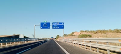 The image shows a view from a vehicle traveling on a highway in Valmojado, Spain. Two blue highway signs are visible on the left side of the road. The first sign indicates E-90, A-5, Maqueda, and Badajoz, with arrows pointing down. The second sign, positioned to the right of the first, shows 500 meters, a highway symbol, and the number 45, followed by CM-41 and A-42 CM-43, with an arrow pointing right. The highway is paved with visible lane markings and is bordered by metal guardrails on both sides. The landscape consists of dry, grassy hills with sparse vegetation and trees under a clear blue sky. In the distance, other vehicles are seen on the highway.