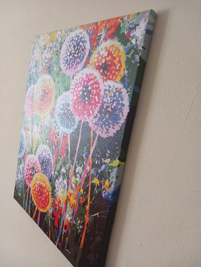 The image shows a colorful painting of dandelions, possibly a paint-by-numbers artwork, mounted on a wall. The painting is predominantly in shades of pink, blue, yellow, and orange, depicting stylized dandelion seed heads against a dark green background. The edges of the canvas are visible, and it appears to be stretched over a frame. The painting is leaning against a textured, light-colored wall, casting a subtle shadow. The composition suggests the artwork is part of an indoor setting, though no other objects or architectural features are clearly visible. The image was taken in Mamaivtsi, Ukraine.