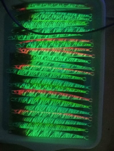 The image shows a tray of 20 fishing lures. The lures are all long and slender, with a green, white, and pink zebra pattern. They are arranged in a neat row, with their heads facing the same direction. The lures are laid in a white plastic tray with raised sides. The tray is lit from above by a fluorescent light. The background is a dark, blurry space. The image was taken in Kebumen, Indonesia, at latitude -7.662, longitude 109.647.