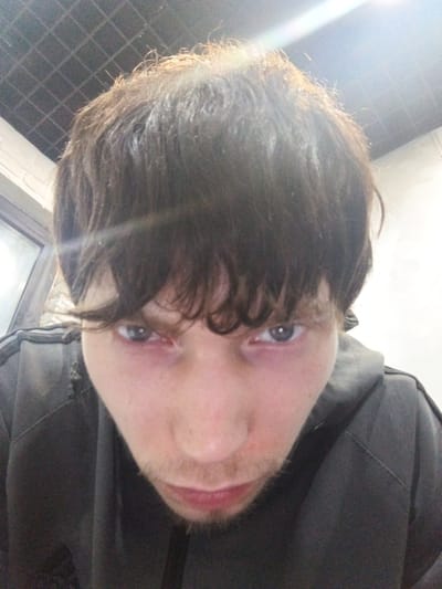A close-up, low-angle selfie shows a young person looking directly at the camera. The subject has medium-length, dark, messy hair that falls across their forehead. Their eyes are open, and they have faint stubble on their chin and upper lip. They are wearing a dark gray hooded jacket. The background appears to be indoors, with a tiled ceiling visible at the top of the frame and a window or glass partition on the left. The lighting is bright, creating a lens flare effect at the top. There is no indication of the city of Khanty-Mansiysk, Russia, in the image.