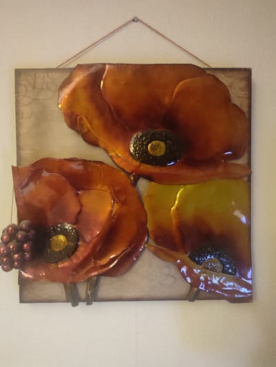 A rectangular, three-dimensional wall artwork is centered in the frame, depicting three stylized flowers resembling poppies and a cluster of grapes. The flowers exhibit a glossy, enameled finish in iridescent shades of orange, red, and yellow. Each flower possesses a dark, textured, raised center with a small yellow accent. The largest flower is positioned at the top, while two smaller flowers are at the bottom. A cluster of approximately ten dark red, spherical objects resembling grapes is affixed to the left side of the lower-left flower. Thin, dark stems are visible beneath the two lower flowers. The artwork's background is a muted beige-brown with subtle, lighter-toned scroll or floral patterns. The piece is suspended from a single metallic hook or nail at the top center by a thin, light-brown cord or string. The artwork is mounted on a plain, light-colored wall. This scene is located in Southampton, United Kingdom.