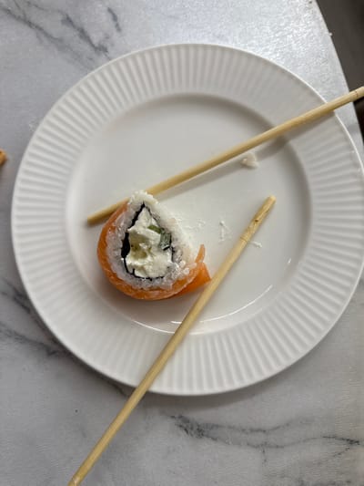 A single sushi roll, featuring salmon, rice, nori, and cream cheese, rests on a white, ribbed plate. Two wooden chopsticks lie diagonally across the plate, one positioned to the left and the other to the right of the sushi. The plate is situated on a marble-patterned surface, suggesting a countertop or table. A small, white, heart-shaped crumb is visible on the plate near the top chopstick. The image is taken from a top-down perspective, with overhead lighting. There is no visible text. The scene appears to be a meal setting, possibly in a home kitchen or dining area in Нижнеяркеево, Russia. The lighting and composition suggest a casual, everyday moment.