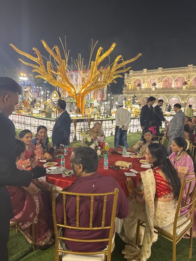 A vibrant outdoor wedding reception is captured at night. Elegantly dressed guests are seated at tables with red tablecloths, enjoying the festivities. A towering, gilded centerpiece resembling abstract branches adds a touch of extravagance. Behind the tables, a beautifully lit building with intricate architectural details serves as a backdrop. The atmosphere is lively and celebratory.