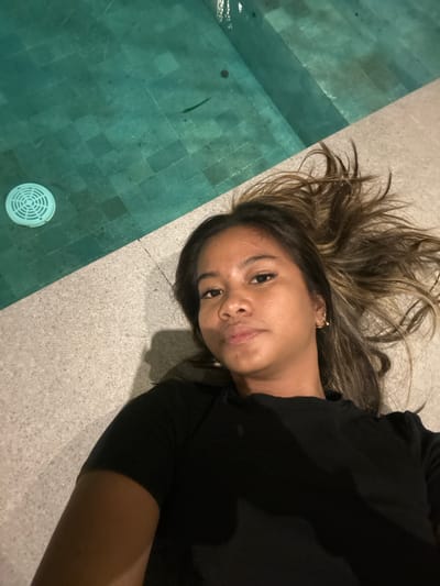 A woman is lying on a stone surface next to a tiled pool. The woman has shoulder-length brown hair with blonde highlights and is wearing a black t-shirt. A circular drain is visible in the pool. The scene appears to be taken outdoors, possibly near a swimming pool in Tumbak Bayuh, Indonesia.