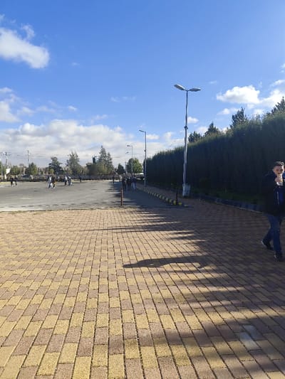 The scene depicts an outdoor urban environment in Jibb as-Safa, Syria, under a clear blue sky with scattered white cumulus clouds. In the foreground, a pathway composed of alternating light brown and yellowish-tan rectangular pavers, arranged in a basket-weave pattern, extends from the bottom of the frame towards the midground. Distinct elongated shadows are cast across this paved area from the right, indicating sunlight from the left.

To the right of the paved pathway, a raised curb with yellow and black striped markings borders a narrow asphalt road or walkway. A tall, silver-colored street light pole with a dual fixture stands prominently along this road. Further down the road, another street light pole is visible. A dense, continuous hedge of dark green foliage runs parallel to the road, forming a barrier.

On the right side of the foreground, a male individual is captured mid-stride, walking leftwards across the paved area. He is wearing a dark jacket, a blue shirt, and blue jeans, and is holding a clear plastic bottle to his mouth, appearing to drink from it.

To the left of the paved pathway, a large, grey asphalt or concrete open area, possibly a plaza or parking lot, is visible. Several groups of individuals are dispersed across this open area, engaged in walking or standing. In the background beyond this open area, additional trees, utility poles, power lines, and distant structures are discernible. Further down the asphalt road to the right, several more individuals are seen walking.