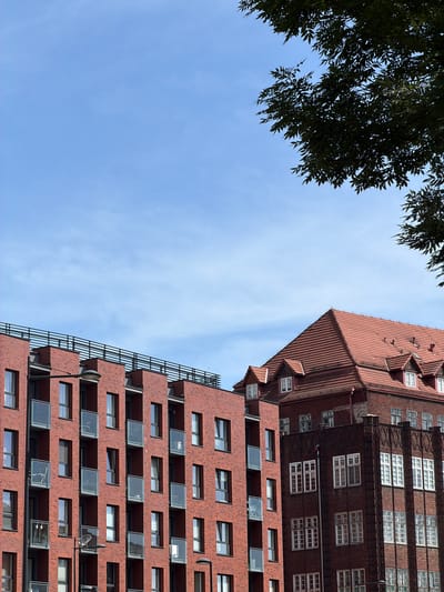 This is a photograph of a bright, modern apartment building made of red brick, with a contrasting older, darker brick building next to it. The older building has a steeply pitched red tile roof and several dormer windows. Both buildings are seen from a low angle, with a clear blue sky and some tree branches in the upper right corner. The apartment building has many windows and balconies with glass railings. There are streetlights visible in front of both buildings. The location is the city of Gdańsk, Poland, specifically near the former shipyard area.