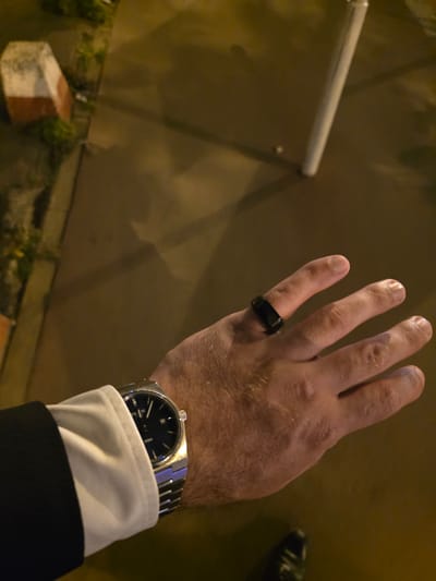 A first-person perspective shot of a person's hand with a black ring on their left ring finger and a silver watch on their left wrist. The hand is positioned over a dark surface, possibly pavement, with a white pillar casting a shadow on the ground. In the background, a white and red marker stone is visible on the left side of the frame. The scene appears to be outdoors at night, with artificial lighting illuminating the area. The location is the city of Montrouge, France.