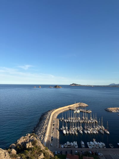 The image shows a high-angle view of a marina in Santa Maria Navarrese, Italy. The marina is filled with numerous sailboats and yachts docked in calm, blue water. A curved concrete and stone breakwater extends into the sea, protecting the harbor. Along the breakwater, cars are parked on a paved road. In the background, a vast expanse of blue sea meets a clear sky, with distant islands and a coastline visible on the horizon. The overall scene depicts a serene coastal landscape with a bustling marina.