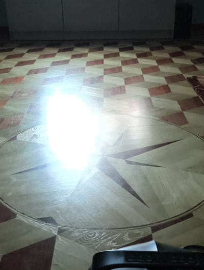 The image shows a floor with a geometric pattern consisting of light and dark brown squares in an optical illusion style, with what appears to be a circular design featuring a starburst pattern at the center. The circular design is bordered by a thin, dark line. A bright glare obscures the center of the starburst pattern. The background shows a wall with cabinetry. Part of an electronic device is visible in the lower right corner. There is no evidence in the image to confirm that this photo was taken in Kolosisty, Russia.