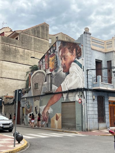 This detailed description pertains to an urban street scene in Picassent, Spain.

The central focus is a two-story building on a street corner, adorned with a large mural. The mural depicts an elderly woman with brown hair tied back, wearing a white shirt with a green collar/stripe. She is shown in profile, looking down and to her right, with her right arm extended downwards and hand open. Behind her, on the upper part of the wall, are painted depictions of various grocery store items, including boxes labeled "Cola" and bottles. The mural continues onto the ground level, showing a hand-crank coffee grinder and a large hand reaching down towards brown cardboard boxes. The signature "CHOMAPICA 2024" is visible in black text on the light blue wall beneath the mural.

The building itself is painted in a light blue-grey color with cream-colored architectural detailing, including horizontal bands and a decorative roofline featuring a small, light-colored statuette or ornament on a pedestal. It has dark brown wooden doors, one of which is open on the upper floor, revealing a dark interior. A small balcony with a black metal railing extends from the upper floor. The ground floor corner features a closed metal roller shutter. Numerous black electrical cables are strung across the building's facade. A circular red and white "no entry" or "stop" sign is attached to the wall below the mural.

To the left of the mural building, a multi-story concrete block building with a muted beige-grey tone is visible, featuring terra cotta roof tiles and an exposed interior structure on one side. A street lamp with a curved black arm extends from this building.

The street level features an asphalt road with a white pedestrian crosswalk. A yellow painted curb separates the road from the reddish-brown tiled sidewalk. Two black spherical bollards are positioned on the sidewalk in the foreground. A grey Peugeot sedan with the license plate "3432 JDK" is partially visible, parked on the left side of the street.

Four young women, wearing shorts and t-shirts/tank tops, are walking away from the viewer on the sidewalk to the right, near the mural. Another person, partially visible on the far right edge, holds a pink object and faces away from the camera. A green palm tree is visible behind the mural building. The sky is overcast with light grey clouds.