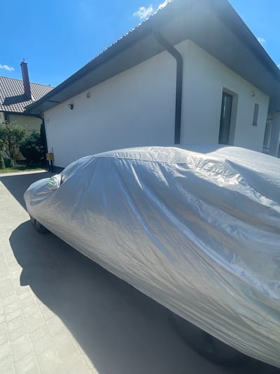 The image shows a car covered with a silver car cover, parked on a paved driveway. The car is partially visible, with its wheel and the draped fabric of the cover taking up most of the frame. The cover is wrinkled and reflects the sunlight, indicating it's a bright day.

To the left of the car, a white building with a dark gutter system is visible. The building has at least two windows, one narrow and one wider. The sky above is clear blue with a few scattered white clouds. The sunlight suggests it is daytime, likely midday.

In the background, a portion of a tiled roof of another building is visible, with a decorative chimney-like structure. Some greenery, possibly trees or bushes, can be seen near the base of the building. The paving stones of the driveway are clearly visible in the foreground and to the right, with a large shadow cast by the covered car. The overall scene is residential and appears to be outdoors. There is no visible text in the image. The location context provided is Мухавецкі сельскі Савет, Belarus.