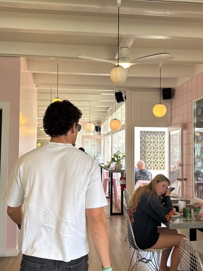 The photograph, taken indoors, shows a man with dark curly hair and sunglasses walking away from the camera. He is wearing a white t-shirt and dark jeans. In the background, a young woman with blonde hair is seated at a table, looking towards the camera while holding a phone to her ear. She wears a dark shirt with a flame design. The setting appears to be a restaurant with pink tiled walls and white ceiling beams. A ceiling fan with a spherical light fixture is visible. Other patrons can be seen in the background. The restaurant setting and the presence of a Heineken bracelet on the man's wrist suggest a casual atmosphere. The location is likely in Palm Desert, United States.