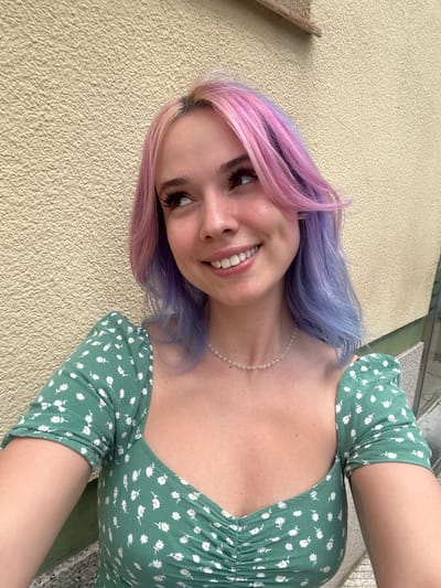 In an outdoor urban setting, a young woman with multi-colored hair, featuring pink and purple tones, smiles and looks to her left. She is wearing a green dress with a white floral pattern and a delicate beaded necklace. The background consists of a textured, pale yellow wall and a hint of greenery, suggesting a location in Oviedo/uviéu, Spain, possibly near a building or park.