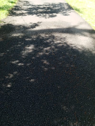 A dark asphalt path is partially shaded by tree leaves above. The shadows are fragmented, creating patterns of light and dark across the rough surface. Green grass edges the path on either side. The asphalt texture is visible in detail, with small pebbles embedded within.