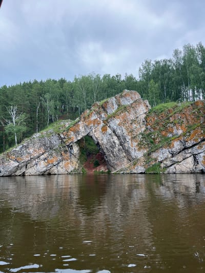 The image depicts a natural landscape featuring a rocky cliff face with an archway, situated along a river. The cliff is composed of layered rocks, exhibiting various shades of gray and orange, with patches of green moss and grass growing on its surface. The archway carved into the rock face reveals a dark, reddish-brown interior, suggesting a cave or recess. The river in the foreground is dark and reflects the overcast sky and the surrounding environment. Behind the cliff, a dense forest of green trees, including birch and pine, stretches upwards towards a cloudy sky. The scene is set in the city of Martyush, Russia, indicating a location within a natural park or scenic area. There are no people or animals visible in the image.