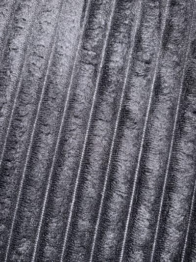 The image is a close-up, overhead shot of a dark gray, ribbed fabric. The ribs are oriented diagonally from the top left to the bottom right of the frame, with a slight shimmer suggesting a synthetic material or a fine, metallic thread woven into the fabric. The texture of the fabric appears plush and somewhat fuzzy, with individual fibers visible within the ridges. The lighting creates shadows and highlights that emphasize the texture and the depth of the ribs. There are no people, actions, or environmental details visible that would suggest a specific location in Evere, Belgium, or any other place. The image is purely an abstract depiction of a textured surface.