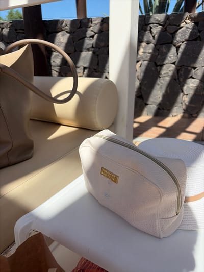 The image shows a close-up of two items on a white surface, likely a table or lounger, in what appears to be an outdoor setting with bright sunlight and shadows. To the left is the lower portion of a taupe leather handbag with a curved handle visible. To the right is a light-colored cosmetic bag with a zippered opening and a gold plaque on the front that reads "LIERAC". Behind the cosmetic bag, a white straw hat with a tan band is partially visible. The background consists of a dark stone wall and a glimpse of blue sky. The overall impression is one of a relaxed, sunny environment, possibly a resort or patio area. While the image does not explicitly indicate Antigua, Spain, the stone wall and sunny ambiance are consistent with the architectural style and climate of the region.