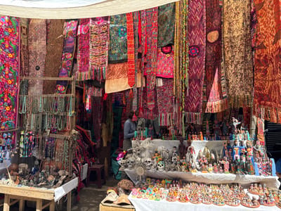 This vibrant image captures a bustling market stall in Chichicastenango, Guatemala, overflowing with a rich array of local crafts and textiles. The scene is dominated by numerous intricately patterned fabrics, scarves, and blankets hanging vertically, creating a colorful backdrop. These textiles display a diverse range of designs, including floral motifs, geometric patterns, and abstract shapes, rendered in bright hues like fuchsia, deep red, purple, green, and earthy tones. Many of the fabrics feature decorative fringed edges.

In the foreground and mid-ground, various merchandise is displayed on wooden tables and shelves. To the left, several racks are laden with long beaded necklaces, primarily in shades of turquoise, green, and brown. Below these, a table exhibits a collection of smaller items, including decorative skulls and other small figurines.

The central and right-hand sections of the stall showcase a wide assortment of pottery and handcrafted figurines. Larger, brightly painted ceramic figures, some depicting people, possibly Mayan or religious figures, are arranged on a raised display. Below them, countless smaller, uniform figurines, often made of clay or ceramic and brightly painted, are meticulously organized in rows on a lower white-covered surface. Some notable crafts include decorated skulls and animal figures.

Behind the central display, partially obscured by merchandise, one person wearing a blue-grey top can be seen attending the stall, suggesting they might be a vendor. The overall impression is one of lively commerce and cultural display.

The lighting indicates it is daytime, likely sunny, as bright light illuminates the colorful goods. A light-colored fabric canopy stretches across the top, providing shade. There is no visible text in the image.