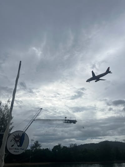 An airplane is in flight against a cloudy sky. Below the airplane, a wake park facility is visible with cables and structures. A sign for "Sport Wake Park Phuket" is also present. The scene appears to be a coastal area with trees and water. The overall impression is one of an outdoor recreational area with an active airport nearby.