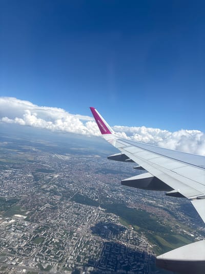 This is a high-angle shot taken from an airplane window, looking down at a sprawling urban landscape and the sky. The dominant subjects are the airplane's wing, which occupies the right side of the frame, and the city below. The wing is a light grey, with visible flaps and structural elements. A portion of the Wizz Air logo, in magenta and white, is visible on the wing's edge.
The scene below is a densely packed city with a mix of buildings, interspersed with patches of green spaces and what appear to be parks or larger recreational areas. Roads and infrastructure are visible, weaving through the urban fabric. The perspective is from a considerable altitude, providing a bird's-eye view of the city's layout and scale.
To the left of the frame, the sky is a vibrant blue, dotted with large, fluffy white cumulus clouds. The clouds create a dramatic contrast with the clear sky, and some appear to be forming cumulonimbus formations.
The time of day appears to be daytime, likely morning or afternoon, given the bright sunlight illuminating the scene and casting shadows. The weather seems fair with good visibility, allowing for a clear view of the city from altitude. The presence of the airplane wing suggests the activity taking place is air travel, with the photo captured during a flight.
Visible text includes "wizzair.com" on the airplane's wing. The location context provided is Bozhurishte, Bulgaria, which would place the urban area seen in the image within or near that region. No people are directly visible in the frame, and the overall mood conveyed is one of observation and perspective, looking down on the world from above.