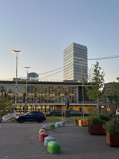 The image depicts a serene urban scene captured during either the golden hour of dawn or dusk, evidenced by the warm glow of artificial lights and the soft, clear sky. The perspective is from street level, looking towards a complex of modern buildings.
In the background, a tall, slender skyscraper with a reflective glass facade stands prominently. The name "Taylor Wessing" is visible in large letters on its side. Below and slightly in front of this skyscraper is a lower, wider building, also featuring extensive glass panels that reflect interior lights. The words "Eindhoven Centraal" are clearly visible on the roofline of this building, indicating it is Eindhoven Central Station or an adjacent structure.
In the mid-ground and foreground, a street or paved area is visible, with a dark-colored car parked on the right. A distinctive line of colorful, rounded bollards – including shades of green, light blue, pink, red, and purple – separates the street from a pedestrian zone or green space. Several streetlights with bright, warm yellow illumination are active, casting light onto the scene. Trees and planters with greenery are interspersed along the street, adding natural elements to the urban landscape. A statue of a figure on a pedestal can be seen near the lower building.
The sky is a clear, light blue, transitioning to a warmer hue near the horizon, consistent with the time of day. Power lines are visible against the sky in the upper left. The scene is devoid of people, contributing to a calm and quiet atmosphere.
