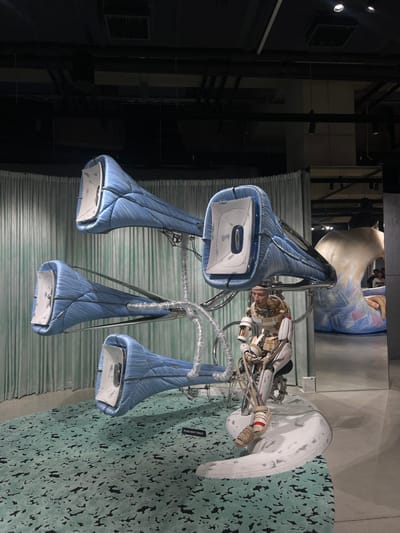 An indoor art installation is depicted within an exhibition space in Seongdong-gu, South Korea. The central subject is a complex apparatus consisting of four large, elongated, horn-like structures wrapped in textured blue material, resembling organic tissue or muscle. These structures, each approximately 1.5 to 2 meters in length, feature reflective, concave white interiors and are mounted at varying angles on a metallic, branching support frame. Numerous cables and tubes interconnect the structures and extend downwards.
Below and integrated with this framework sits a humanoid figure, likely a mannequin or sculpture, dressed in a partial, skeletal-style exosuit. The figure wears a helmet-like headpiece that encases the head and neck, revealing a human-like face within. Its posture is seated, with mechanical components forming parts of its body and limbs. The figure rests on a white, undulating sculptural base that emerges from the floor.
The immediate floor area beneath the installation is covered by a light green carpet with an irregular dark green/black abstract pattern. A small black sign with white text, "PLEASE DO NOT TOUCH," is positioned on this carpet. The background to the left features a light green, vertically pleated curtain. To the right, a large reflective surface (mirror or polished panel) reveals a distorted reflection of a substantial, organic, flesh-toned sculpture, along with blurred figures of two individuals in the deeper background. The ceiling is dark, exposing black industrial ducts, pipes, and recessed lighting fixtures. No active interactions or dynamic movements are present in the foreground scene.