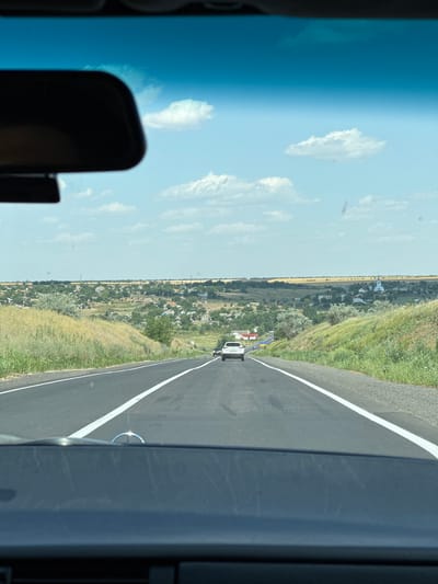 The image is taken from the interior of a car, looking out through the windshield at a paved road stretching into the distance. A white sedan is visible ahead on the road. On either side of the road, there are grassy slopes. In the background, a rural landscape unfolds, characterized by scattered houses, trees, and fields under a partly cloudy sky. There is a distinct white church building with a blue dome visible in the middle-ground of the settlement. The scene depicts a journey through a Ukrainian countryside, likely in or near the city of Kalynivka, Ukraine, given the architecture and landscape typical of the region.