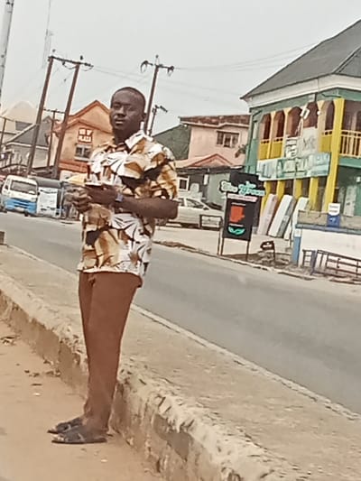The image captures an adult male standing on a dirt and sandy surface next to a concrete curb on a street in Orie Uratta, Nigeria. The sky is overcast. The man, appearing to be of African descent, wears a short-sleeved collared shirt with a brown, white, and black leaf-like pattern, brown trousers, and dark sandals. A dark watch is visible on his left wrist. He is holding a dark-colored smartphone horizontally with both hands at waist level.
Behind the subject is a grey paved road. On the left side of the road, a white and blue minibus is parked, and a lighter-colored car is visible further down. Buildings line both sides of the street. On the left, a light-colored building with "OY PLAZA" inscribed on its facade is visible, along with other residential structures, including one with a reddish roof. Several wooden utility poles with multiple overhead wires span the background. On the right side of the road, a prominent multi-story building features a dark sloped roof, a teal-green lower facade, and yellow columns supporting an upper balcony. Adjacent to this building, several mattress-like objects are stacked or leaning against the roadside, and a black A-frame sign with "Bio Green" and a leaf logo is present.