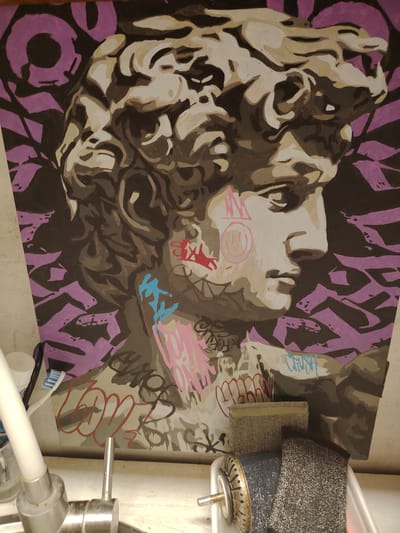 The media file contains a painting of Michelangelo's David bust, rendered in a stylized, graphic manner with a limited color palette of grays, whites, blacks, and purples. The background features abstract, angular purple shapes. The painting is adorned with graffiti tags in various colors, including pink, blue, red, and white, applied over the bust and background. Some of the graffiti is abstract, while other tags appear to be stylized lettering.
The bust itself is depicted in profile, facing right, with attention to the sculpted hair and facial features. The graffiti creates a juxtaposition between the classical art form and contemporary street art. Visible graffiti tags include what appear to be the words "LOVE" in red on the lower left, and "CRUSH" in blue on the right side of the bust. There are also circular shapes with smiley faces or symbols drawn in pink.
The painting is presented in a setting that suggests a sink or countertop area, with a toothbrush and a metal cleaning tool partially visible in the foreground. These objects, along with the overall lighting, imply an indoor, possibly domestic or workshop environment. The lighting is somewhat diffused, suggesting it is not direct sunlight. No specific time of day or weather conditions can be determined from the image. The overall mood conveyed is one of artistic fusion and urban expression.