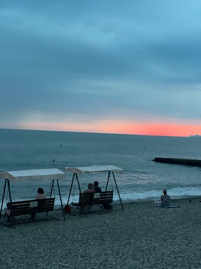 The image depicts a serene evening scene on a pebble beach in Khostinskiy, Russia. The sky displays a gradient of soft blues and pinks, indicative of a sunset, with the horizon clearly defined by the meeting of the sky and the calm sea. Two beach swings, each with a white canopy, are positioned on the shore. On the left swing, a person is seated facing the sea. On the right swing, a couple is seated together, also facing the sea, with their arms around each other. To the right, a lone individual is sitting on a mat on the beach, looking towards the sea and holding a phone. Gentle waves lap the shore, and a dark, man-made pier or breakwater extends into the sea on the right side of the frame. The beach itself is covered in smooth, dark grey pebbles.
