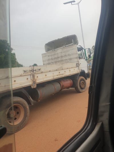 A flatbed truck is shown from a side-view perspective, partially obscured by the interior of a vehicle. The truck is white with a weathered wooden flatbed and a white cab. Two large cylindrical tanks, one appearing red and the other gray, are secured underneath the flatbed. The truck is on a reddish-brown dirt road. In the background, a tall lamppost with a modern, rectangular fixture stands against an overcast sky. There are power lines overhead and some greenery, including trees, visible to the left and right. The scene is likely outdoors in a somewhat rural or suburban area.