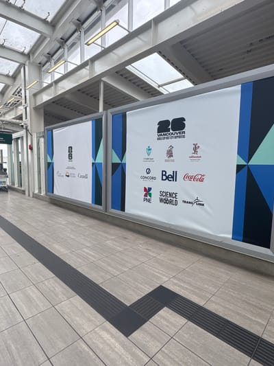 The image shows a section of an outdoor walkway in Richmond, Canada, likely at a transit station given the presence of tactile paving on the ground. Two large banners are displayed on the wall of the walkway. The banners feature a geometric design in shades of blue, black, and green at the top.

The upper banner prominently displays the text "VANCOUVER WORLD CUP HOST CITY SUPPORTERS" along with logos of various sponsors and supporting organizations. These include "x"mək əyəm MUSQUEAM INDIAN BAND", "səlilwətat UAMISH NATION", "Bell", "Coca-Cola", "SCIENCE WORLD", "TRANS LINK", and "PNE".

The lower banner also displays a similar geometric design and features the text "Canada", "BRITISH COLUMBIA", and "VANCOUVER". It also includes what appears to be a government crest and possibly more sponsor logos that are partially obscured or not clearly legible.

The walkway is paved with light-colored tiles, and a dark-colored tactile paving strip runs along the edge of the walkway, indicating a potential hazard or change in surface. The ceiling above appears to be constructed with metal beams and ribbed panels, with fluorescent lighting fixtures visible. The overall impression is of a clean, modern public space that is decorated to commemorate a significant event, likely the World Cup. The lighting suggests it could be daytime, but the weather appears overcast. There are no people visible in the immediate frame.