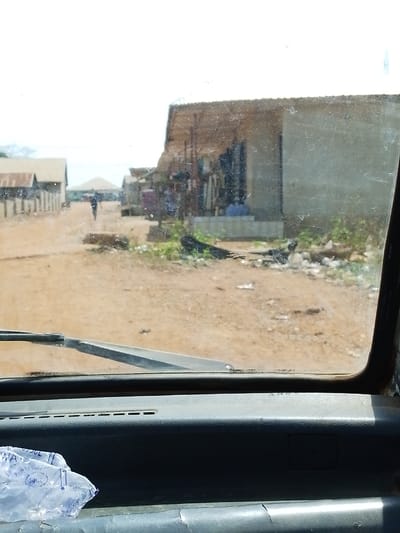 The image presents a street scene in Oke Onigbin, Nigeria, viewed from within a vehicle through a smudged and scratched windshield. The foreground shows the vehicle's dark grey dashboard, with a single windshield wiper blade positioned horizontally at its base. A crumpled clear plastic bag, partially obscuring the word "WATER" and showing "50CL," rests on the left side of the dashboard.
The exterior scene features a wide, unpaved dirt road, stretching into the distance. Along the immediate right side of the road, a light-colored building with an extended corrugated metal roof forms a covered stall or shop area, displaying various indistinct items. A single individual is seated beneath this covered section. Debris is scattered along the road's edge in front of the structure. Further down the road on the left, two additional single-story buildings with corrugated metal roofs are visible behind a low wall. A lone individual, silhouetted, walks away from the viewer down the dirt road in the mid-ground. The sky above is bright white and hazy, suggesting strong daylight, with sparse vegetation visible at the road's periphery.