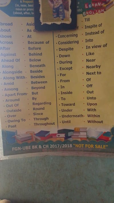 The image is a close-up, slightly angled shot of a yellow poster displaying lists of English words, likely prepositions or conjunctions, categorized by columns. At the top left, a small portion of another image or poster is visible, showing a person's legs in blue jeans and striped socks. To the right of this, the words "LEARN ENGLISH" are prominently displayed in red and blue lettering. The main body of the poster is filled with bullet-pointed lists of words such as "Abroad," "About," "Across," "After," "Against," "Ahead Of," "Along," "Alongside," "Among," "Apart From," "Around," "Out Of," "Outside," "Over," "Owing To," and "Past." These are arranged in multiple columns, with headings or categories that are partially obscured or missing.
A section of text at the top reads "A Preposit (in, near, besi noun or prono (about, after, be". At the bottom of the poster, a dark blue banner contains white text: "FGN-UBE BK & CH 2017/2018 'NOT FOR SALE'". The poster appears to be laminated or printed on a glossy material. The background is a textured, greenish-blue surface, possibly a wall or a table, with faint scratch marks. The lighting suggests an indoor environment, with the light source likely from the front and slightly to the left. The overall impression is educational material, possibly displayed in a classroom or an office setting in Nigeria, given the text at the bottom. No specific time of day, weather, or emotions are discernible.