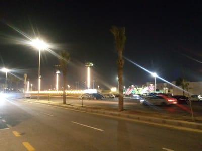 The image presents a night-time street scene in Al Khobar, Saudi Arabia, characterized by an abundance of artificial illumination. The dark sky highlights the bright streetlights, which emit prominent star-like flares and illuminate the asphalt road below. The road features clear white and yellow lane markings, indicating vehicular traffic.
Numerous palm trees are scattered throughout the scene, lining the roadside and appearing in the background. Several cars are visible, both parked and potentially in motion, with their headlights and taillights creating streaks of light across the frame. Along the side of the road, a decorative barrier or fence is adorned with a continuous wavy pattern of golden-yellow lights, adding to the festive atmosphere.
In the mid-ground, a colorful structure resembling an inflatable bouncy castle or amusement ride is brightly lit in various hues including green, pink, and blue, suggesting a recreational area or event. The scene depicts general evening activity in an urban or suburban setting. The time of day is clearly night, and the weather appears clear. No visible text is present in the image.